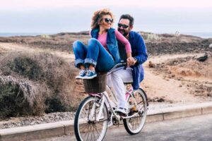 A woman in blue jeans sits on the handlebars of a bicycle ridden by a smiling man