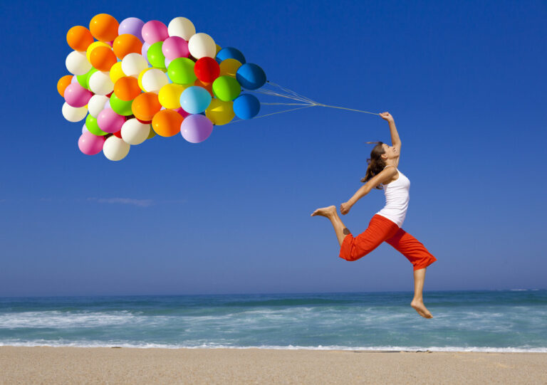 Energetic woman jumping on a sandy beach while holding a large bunch of colorful balloons