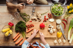An overhead shot of a wooden kitchen table where someone is slicing fresh vegetables (peppers, carrots, mushrooms) on a cutting board, alongside salads, herbs, and wine