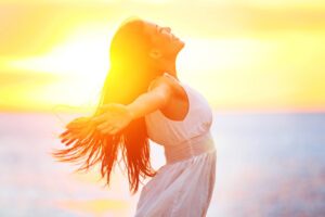 A woman in a white dress with her arms outstretched, soaking in bright, warm sunlight from above