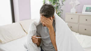 Man in bed wrapped in a blanket holding a thermometer and touching his forehead