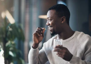 Smiling man in a cream sweater holding a glass of water and preparing to take a vitamin capsule