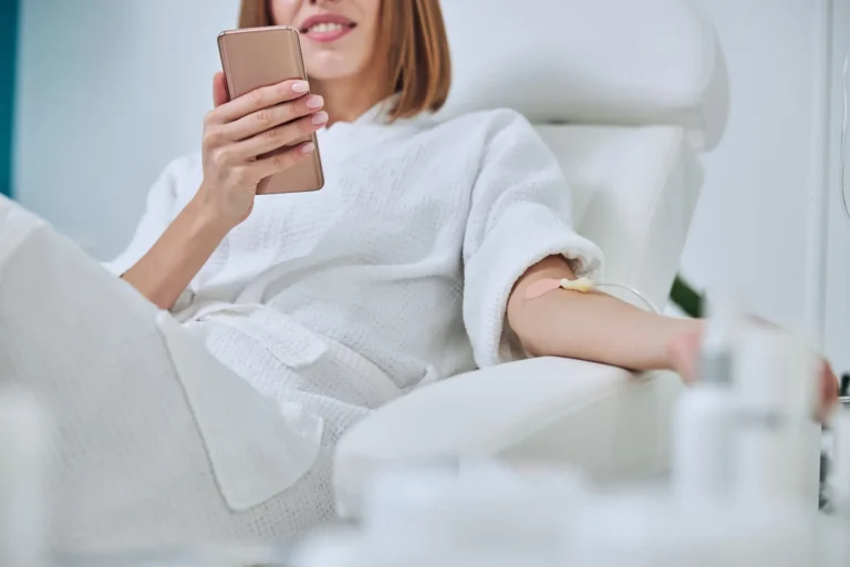 Woman in a white robe using a smartphone while receiving an IV therapy treatment in her arm