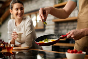 Person cooking eggs while another enjoys breakfast in a cozy kitchen