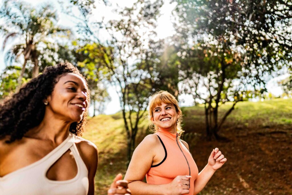 Two women jogging and smiling together on a tree-lined path in a park