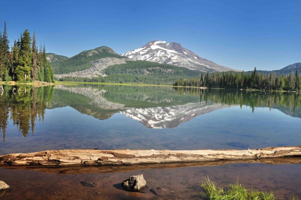 Snow-capped mountain reflected in a calm alpine lake surrounded by evergreen trees under a clear blue sky