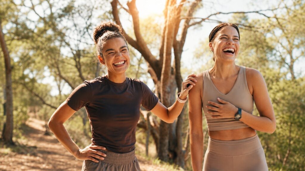 Two smiling women laugh together outdoors in a sunlit forest after a workout, representing vitality, confidence, and healthy aging. Warm natural light and trees create an inviting wellness setting. Featured image for Flow Wellness, a medical wellness clinic providing bioidentical hormone replacement therapy (BHRT), menopause care, and hormone optimization in Oregon and Washington.