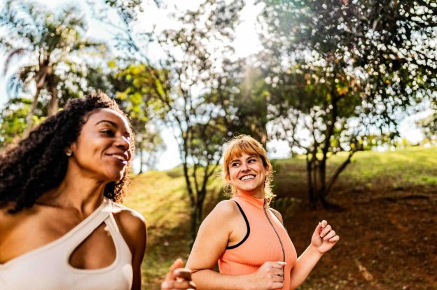 Two women jogging and smiling together on a tree-lined path in a park