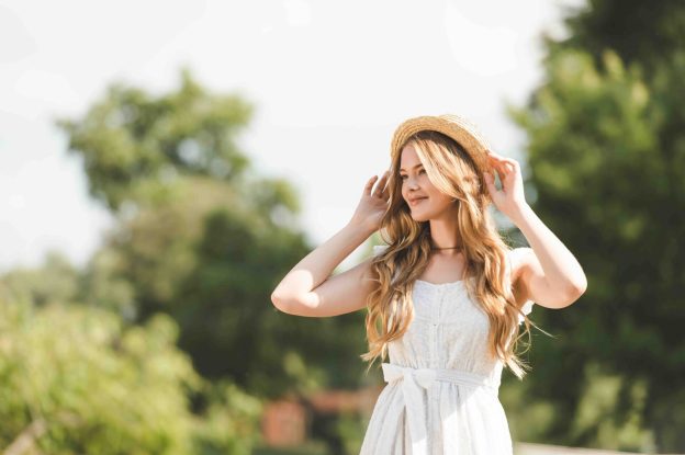Young woman in a white dress and straw hat standing outdoors in sunlight with trees in the background